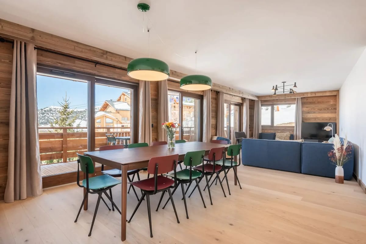 Modern dining and living area with large windows, wooden walls, colorful chairs, a long table, and a sofa. Scenic mountain view outside.