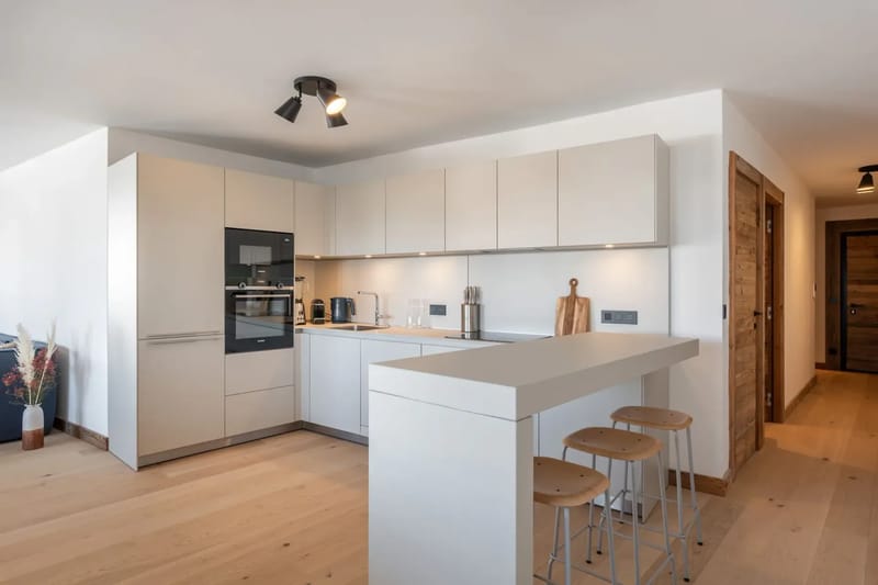 Cocina moderna con armarios blancos, un horno integrado, suelo de madera clara, una pequeña zona de barra con taburetes e iluminación en el techo.