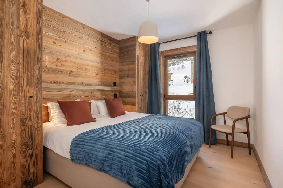 Cozy bedroom with a large bed, blue blanket, brown pillows, wooden accent wall, window with blue curtains, and a wooden chair