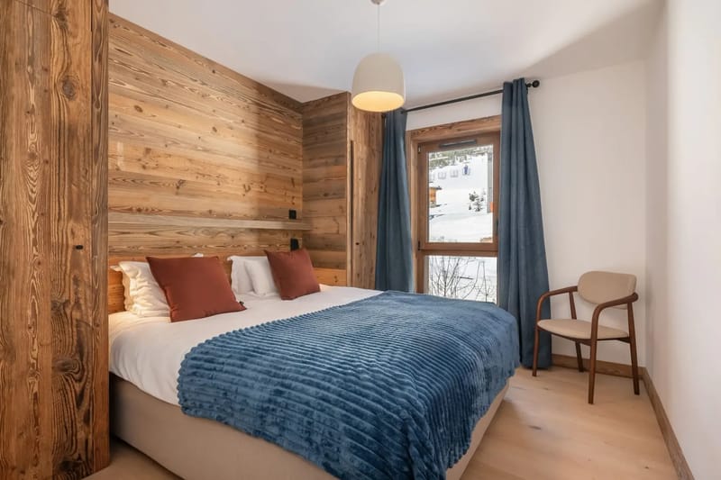 Dormitorio acogedor con una cama grande, una manta azul, almohadas marrones, una pared decorativa de madera, una ventana con cortinas azules y una silla de madera