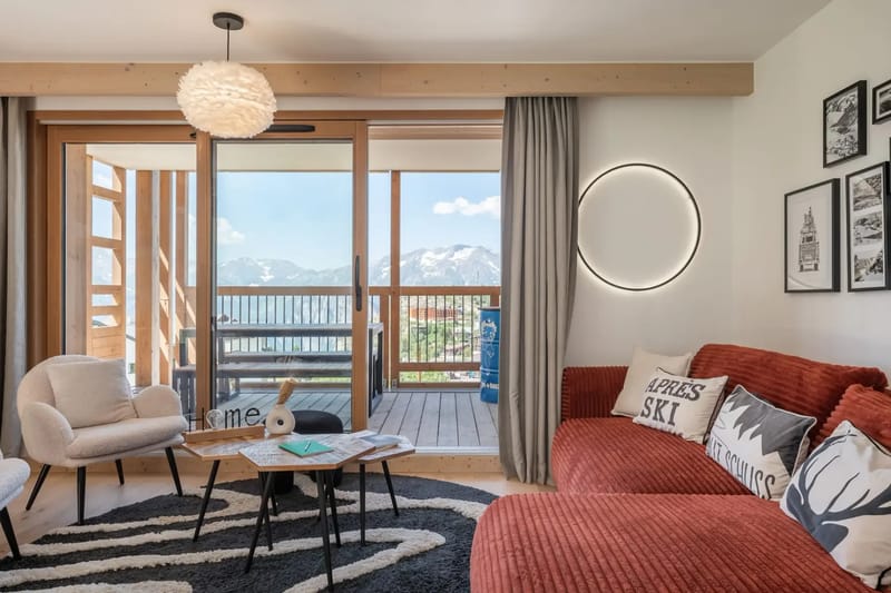 Modern living room with red sofa, white armchairs, a zebra-patterned rug, artwork, and glass doors leading to a balcony with mountain views.