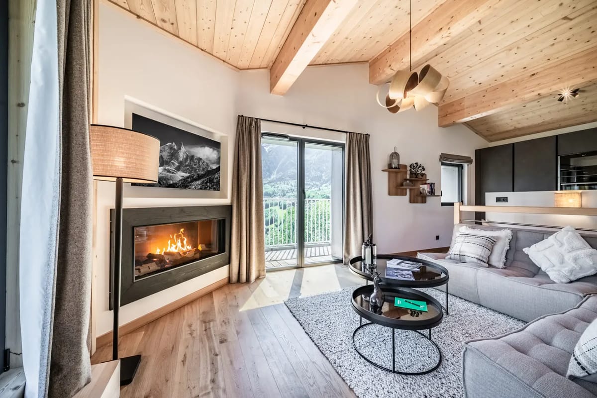 Cozy living room with wooden ceiling, plush sectional sofa, modern fireplace, round coffee table, and large window overlooking a scenic view.