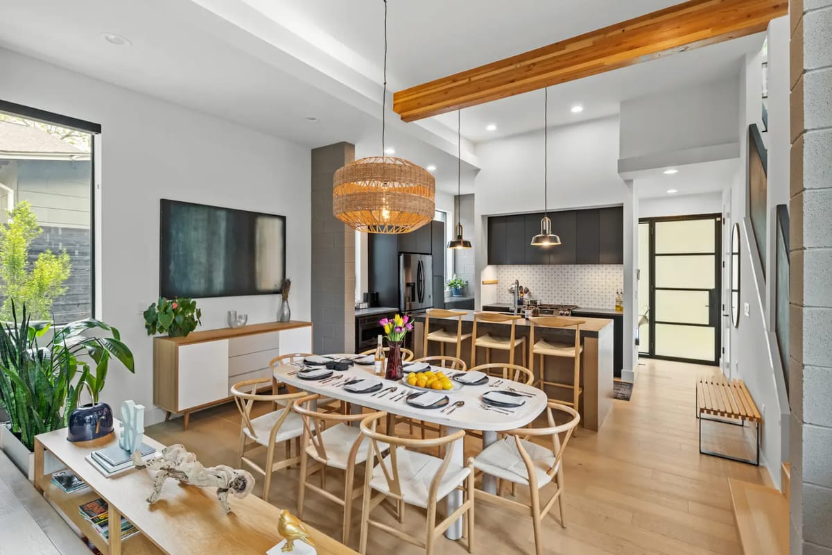 Modern open-concept dining and kitchen area with a wooden table set, wicker light fixture, bar seating, and sleek cabinetry, accented by greenery.