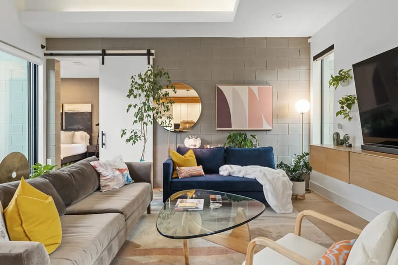 Sala de estar moderna con dos sofás, plantas, un televisor grande, detalles de madera y un comedor abierto. Iluminación brillante y decoración contemporánea.