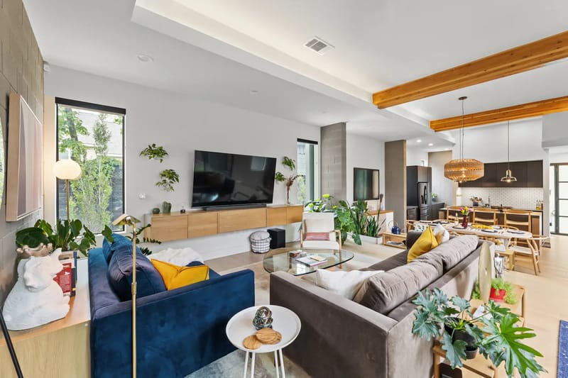 Sala de estar moderna con dos sofás, plantas, un televisor grande, detalles de madera y un comedor abierto. Iluminación brillante y decoración contemporánea.
