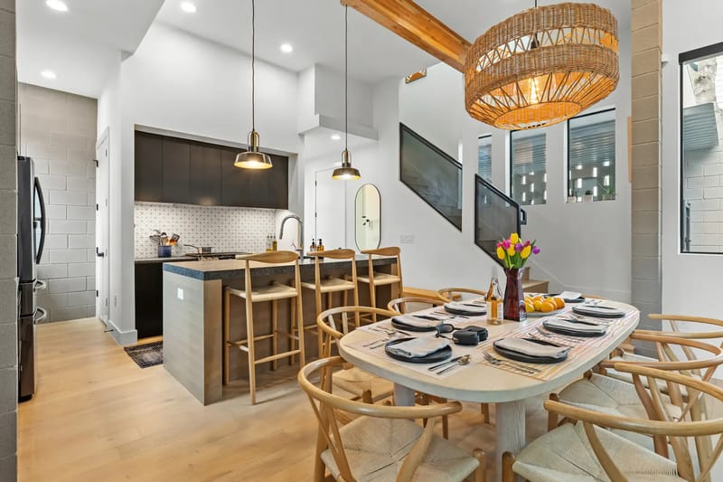 Área moderna de comedor y cocina de concepto abierto, con un conjunto de mesa de madera y sillas, una lámpara de mimbre, asientos en la barra y gabinetes de líneas elegantes, con plantas como detalles decorativos.