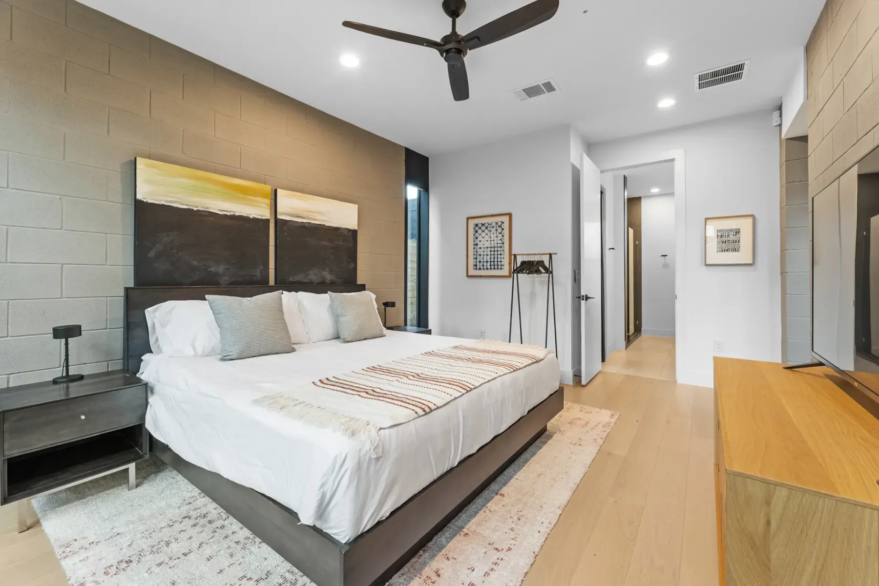 Dormitorio moderno con una cama grande, arte abstracto, ventilador de techo y decoración minimalista. Los pisos de madera clara y los tonos neutros crean un ambiente sereno.