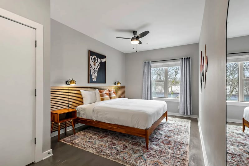 Dormitorio moderno con cama de madera, cabecero a rayas, alfombra estampada y una obra de arte de un cráneo de venado; las ventanas grandes y el ventilador de techo realzan el espacio.