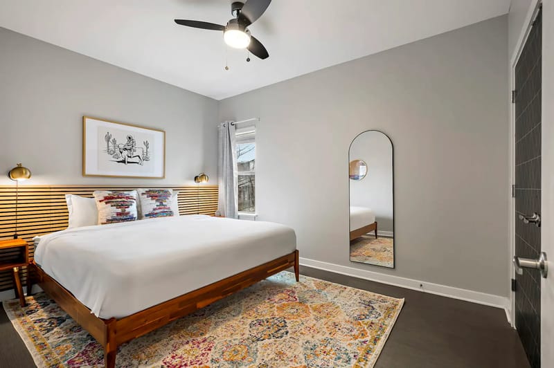 Dormitorio moderno con una cama grande, cabecero de madera, dos lámparas de noche, alfombra colorida, arte en la pared, ventilador de techo y un espejo arqueado.