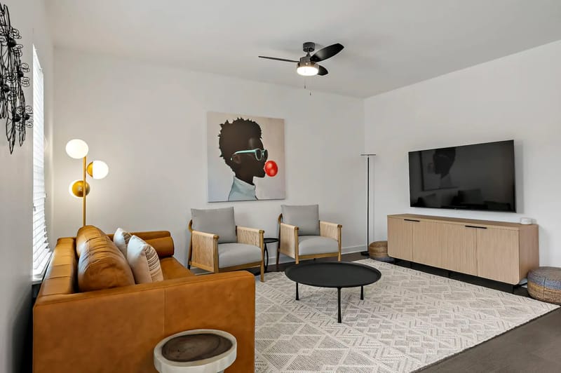 Sala de estar moderna con sofá de cuero marrón, dos sillones, una obra de arte de gran tamaño en la pared, televisor de pantalla plana, ventilador de techo y lámpara de pie decorativa.
