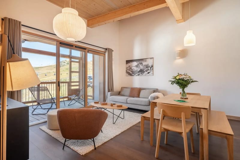 Salón moderno con techo de madera, sofá, sillón, mesa de comedor y acceso al balcón.