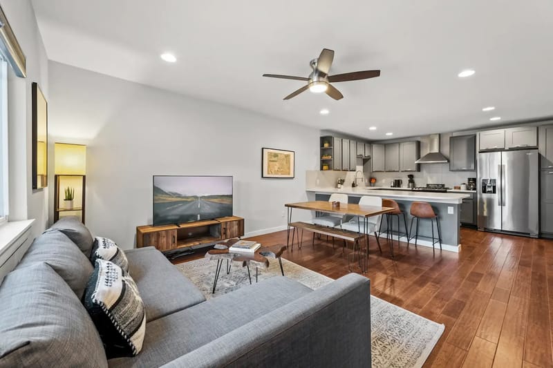 Modern open-plan living room and kitchen with a gray sofa, large TV, wooden dining table, and stainless steel appliances.