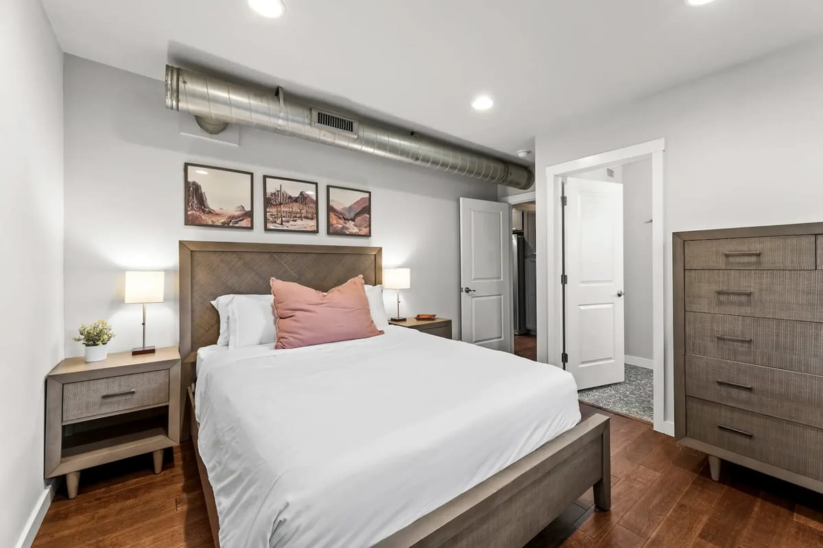Modern bedroom with a double bed, white linens, wooden furniture, and artwork.