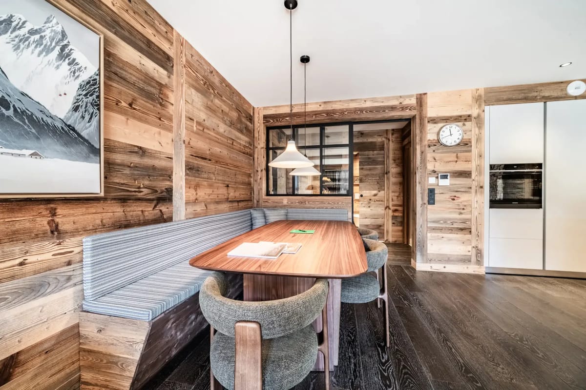 Rustic dining area with wooden walls, a wooden table, green upholstered chairs, a bench, mountain artwork, and a modern light fixture.
