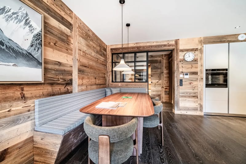 Zona de comedor rústica con paredes de madera, una mesa de madera, sillas tapizadas en verde, un banco, una obra de arte de temática montañosa y una lámpara moderna.