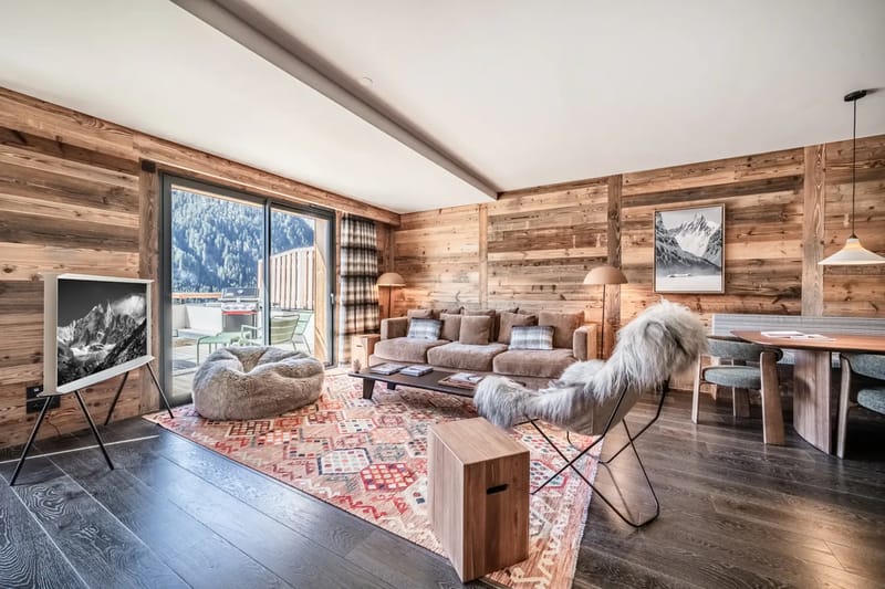 Acogedora sala de estar con paredes de madera, asientos mullidos y una gran ventana con vistas a un pintoresco paisaje de montaña.