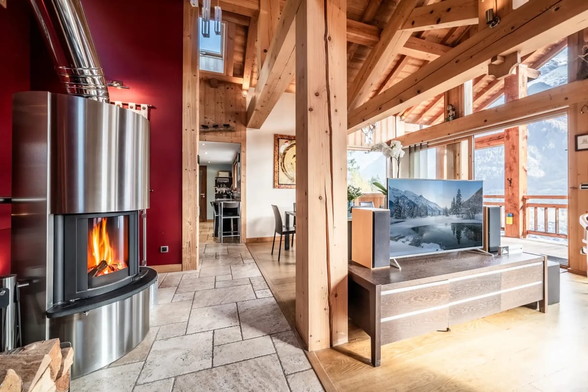 Interior de un acogedor chalet de montaña con una estufa de leña, vigas de madera, suelos de baldosas y un televisor de pantalla plana que muestra un paisaje nevado.