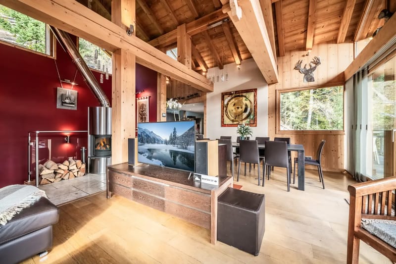 Un salón moderno de planta abierta con vigas de madera, un televisor grande, una chimenea, muebles de comedor y grandes ventanas.