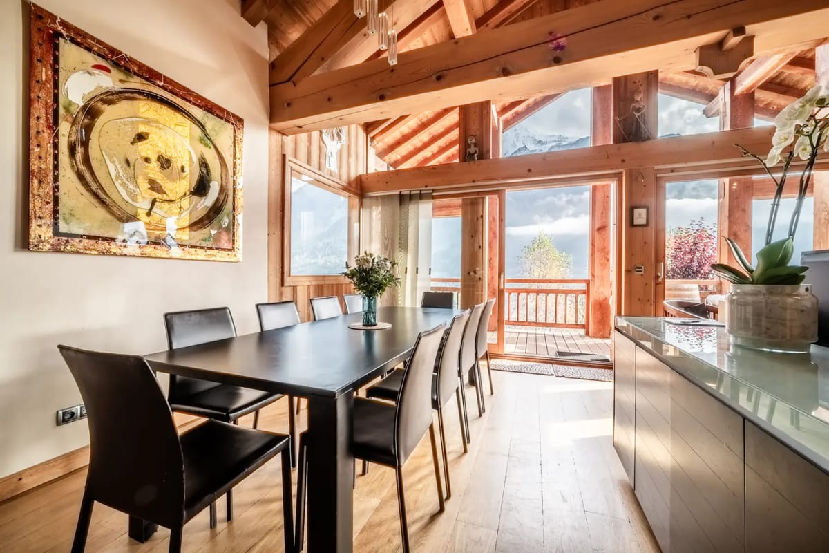 Un comedor con una mesa grande, sillas, arte moderno, vigas de madera, grandes ventanas y vistas a las montañas.