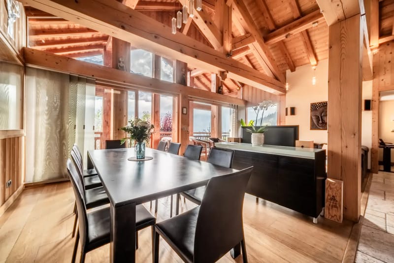 Un comedor espacioso con una mesa de madera y ocho sillas, grandes ventanas y vigas de madera vistas.