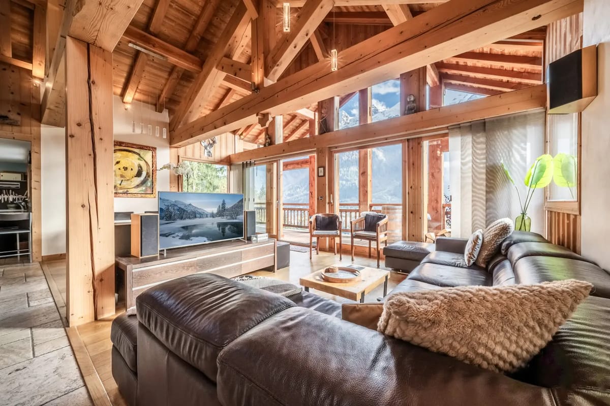 Un acogedor salón con vigas de madera, grandes ventanas con vistas a las montañas, un sofá modular de cuero, dos sillas, un televisor y una mesa de centro cubierta con una alfombra.