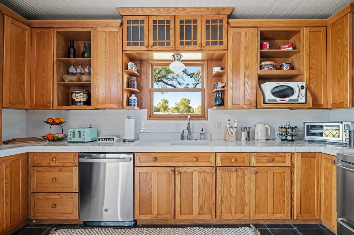 Cocina cálida con acabados de madera, electrodomésticos modernos, una ventana central, estantes, vajilla y decoración.