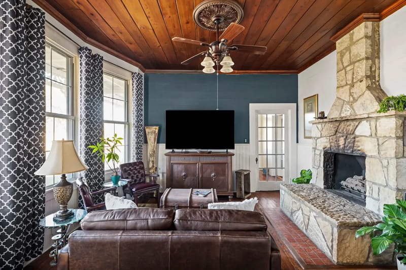 Acogedora sala de estar con un sofá de cuero marrón, una chimenea de piedra, un televisor, un techo de madera y ventanas grandes.
