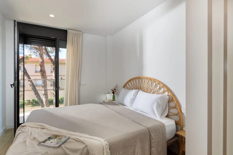 Minimalist bedroom with a double bed, wicker headboard, neutral bedding, and a large window.