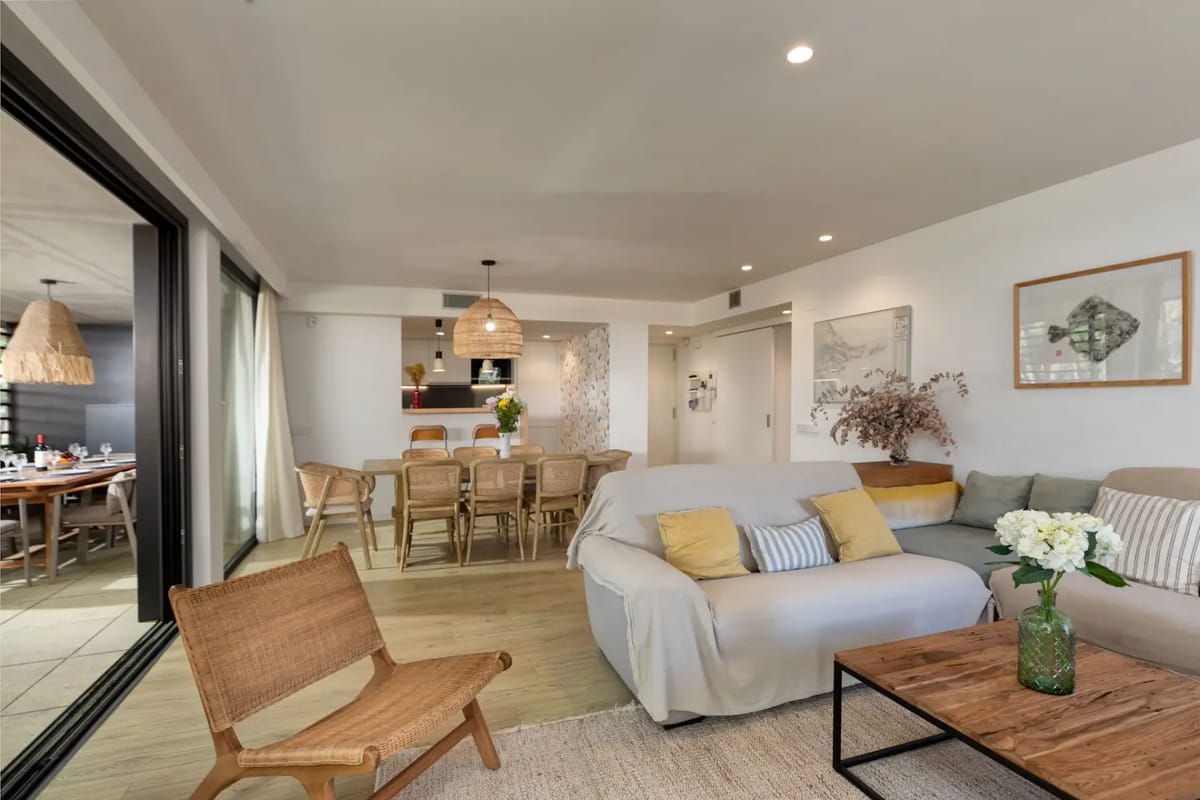 Modern living room with a gray sectional sofa, green pillows, wall-mounted TV, large mirror, nested wooden tables, and sheer curtains covering a bright window.