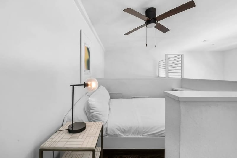 Dormitorio blanco minimalista con ventilador de techo, una mesa auxiliar con una lámpara moderna y una cama junto a la pared.