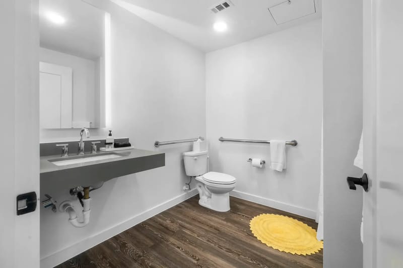 Baño moderno con lavabo suspendido, espejo redondo, inodoro, barras de apoyo y una alfombra amarilla sobre piso de madera.