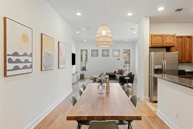 Modern dining room with a wooden table, green chairs, abstract art on white walls, wicker pendant light, and open kitchen area with stainless appliances.