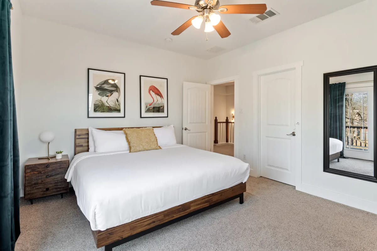 Modern bedroom with a large bed, wooden nightstand, ceiling fan, and art on the wall. A mirror and open door reveal a well-lit hallway.