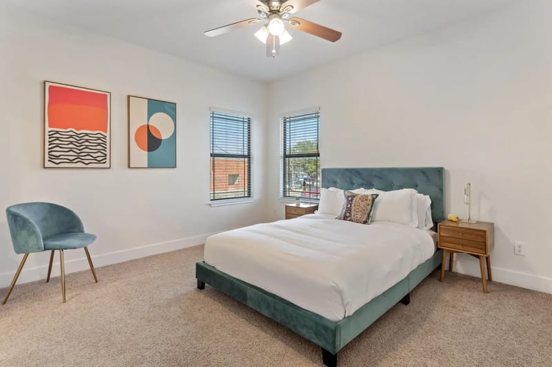 Modern bedroom with a teal bed, white bedding, wooden nightstands, abstract art on walls, a blue chair, carpeted floor, and a ceiling fan.