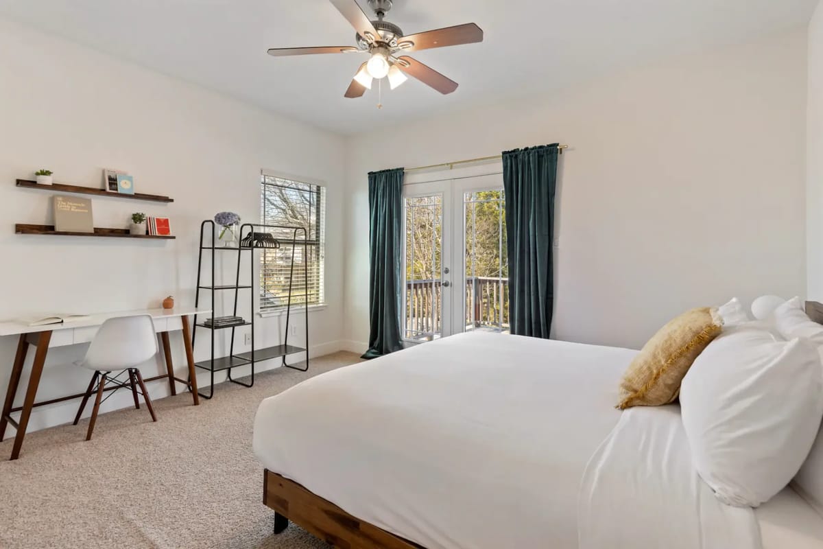 Bright bedroom with a bed, white bedding, desk, chair, shelving, ceiling fan, and large window with dark curtains leading to a balcony.