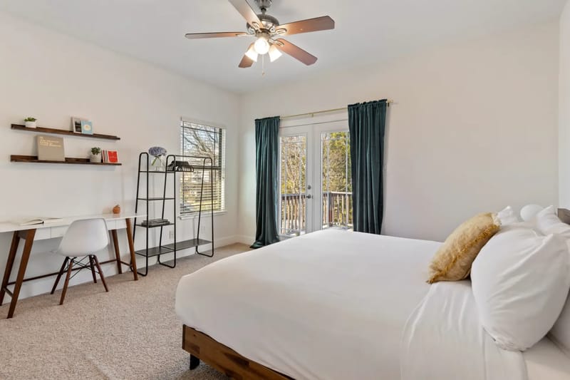 Bright bedroom with a bed, white bedding, desk, chair, shelving, ceiling fan, and large window with dark curtains leading to a balcony.