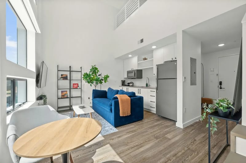 Moderna sala de estar de planta abierta con sofá azul, cocineta, mesa redonda y ventanas grandes.