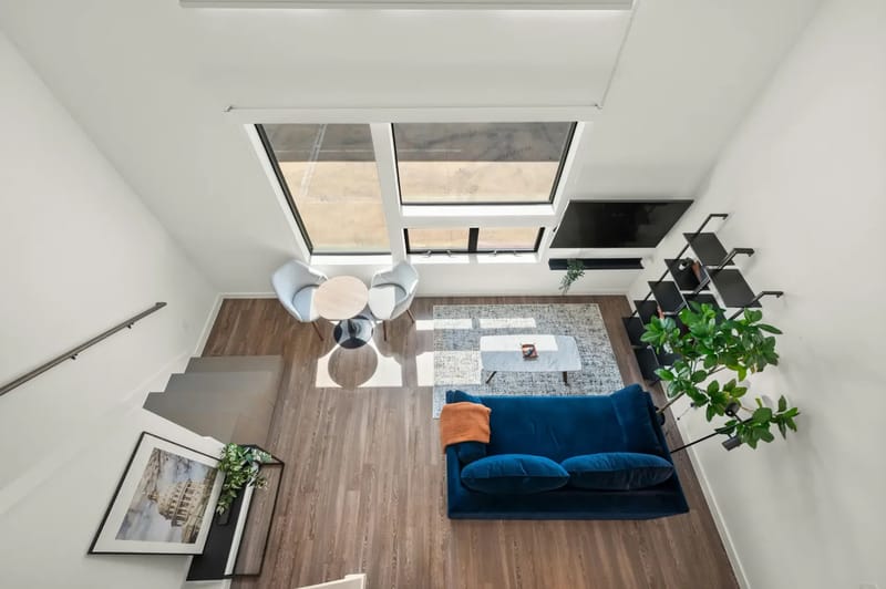 Moderna sala de estar de planta abierta con sofá azul, cocineta, mesa redonda y ventanas grandes.