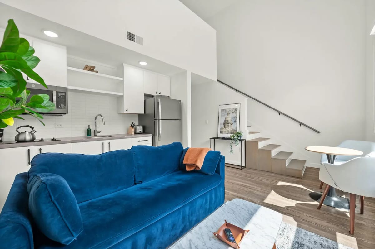 Modern open-plan living area with blue sofa, kitchenette, round table, and large windows.