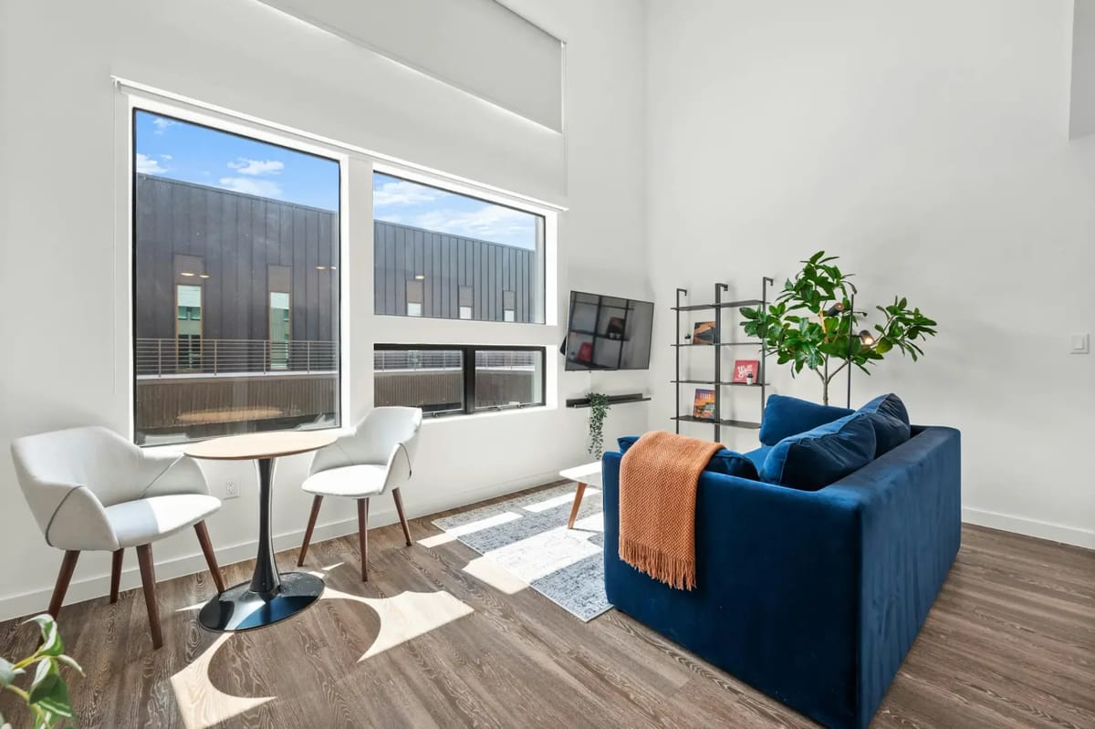 Modern open-plan living area with blue sofa, kitchenette, round table, and large windows.
