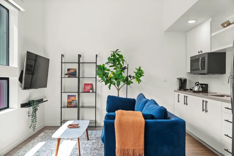 Moderna sala de estar de planta abierta con sofá azul, cocineta, mesa redonda y ventanas grandes.