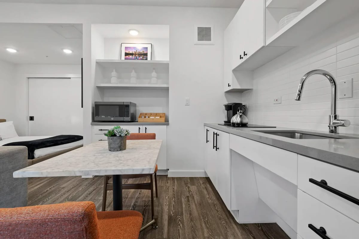 Cocina pequeña con gabinetes blancos, fregadero, microondas y mesa de comedor.