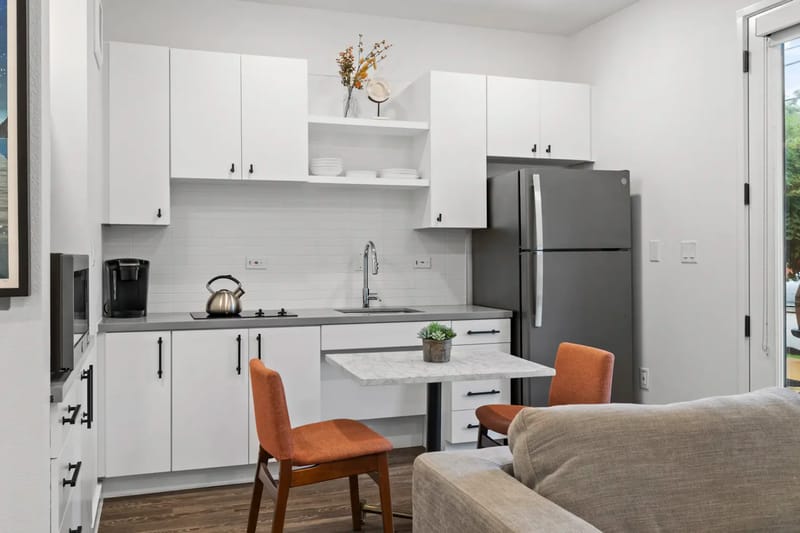 Cocina pequeña moderna con gabinetes blancos, cubierta gris, refrigerador, mesa de comedor y asientos.