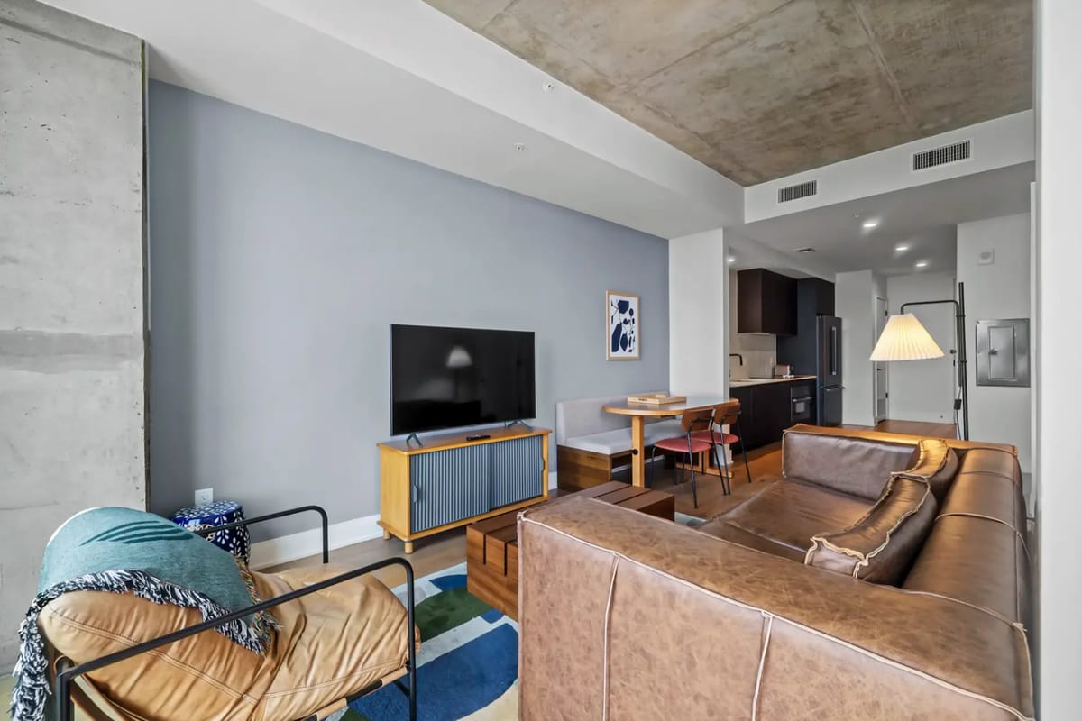 Modern living room with a brown leather sofa, TV on a wooden stand, dining table, and open kitchen in the background.