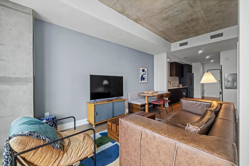 Sala de estar moderna con sofá de cuero marrón, televisor sobre un mueble de madera, mesa de comedor y cocina abierta al fondo.