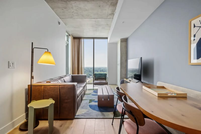 Sala de estar moderna con sofá de cuero marrón, lámpara de pie, mesa de centro de madera y televisor. Una ventana grande ofrece una vista del exterior.