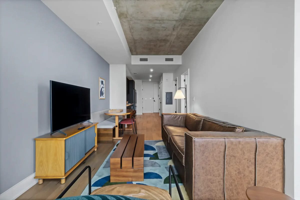 Modern living room with a brown leather sectional, flat-screen TV on a wooden stand, colorful rug, and wooden accents, leading to a hallway.