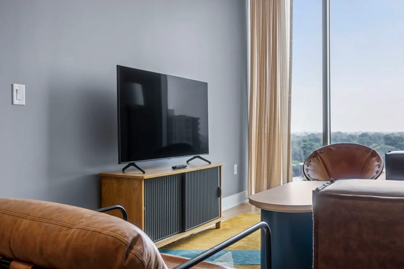Sala de estar con televisor, sillas de cuero beige, mesa redonda y ventanas grandes.