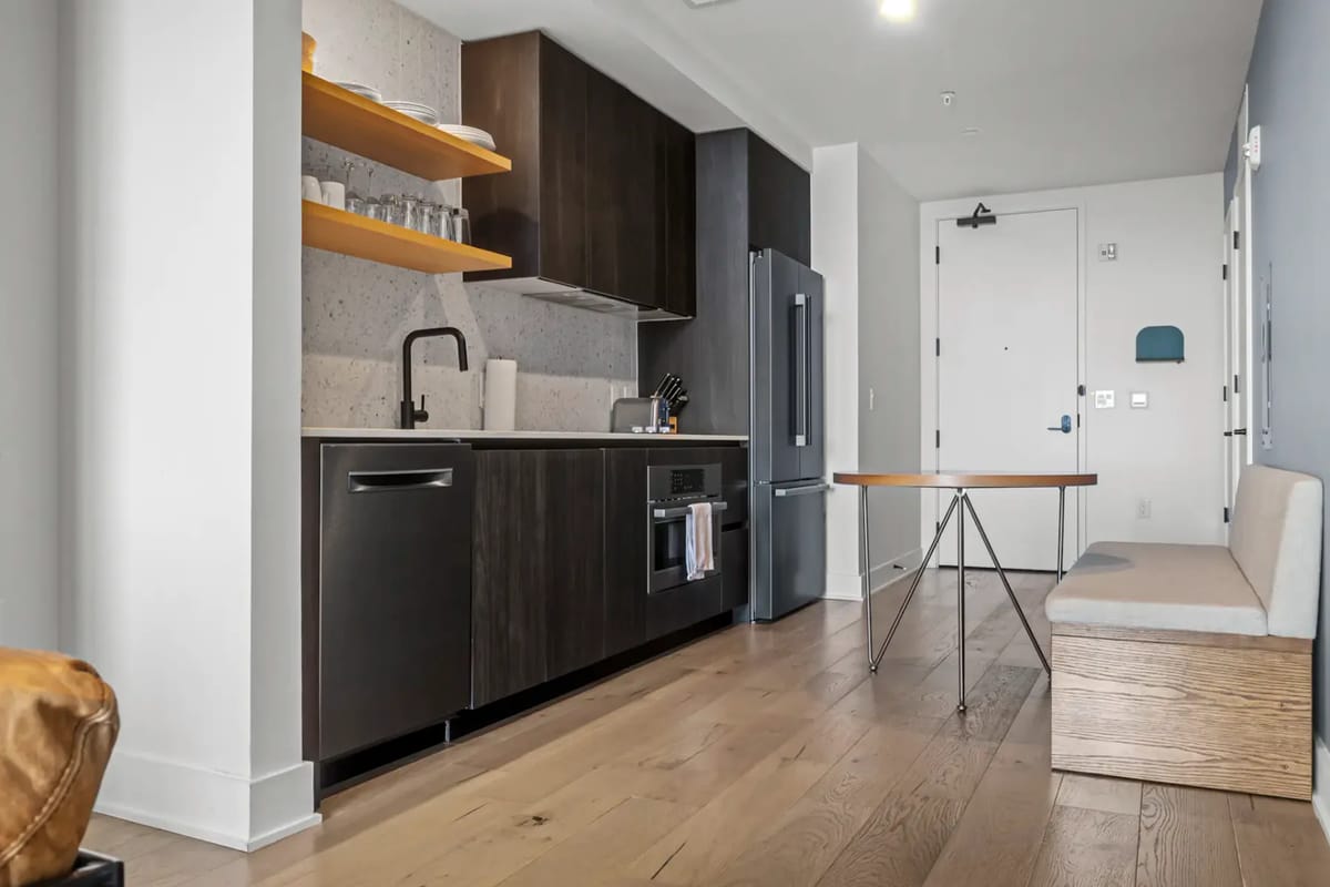 Cocina pequeña moderna con gabinetes oscuros, electrodomésticos de acero inoxidable, estantes y una pequeña zona de comedor.