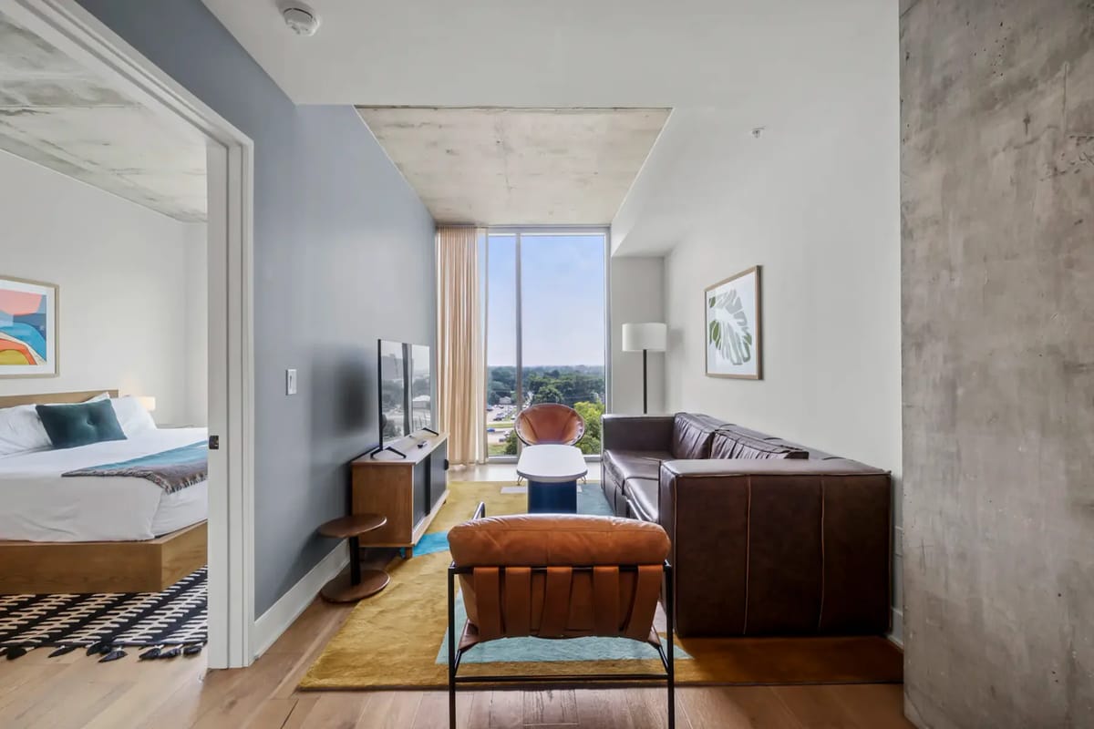 Sala de estar de un apartamento moderno con sofá marrón, televisor y ventana grande; se ve el dormitorio a través de una puerta abierta.
